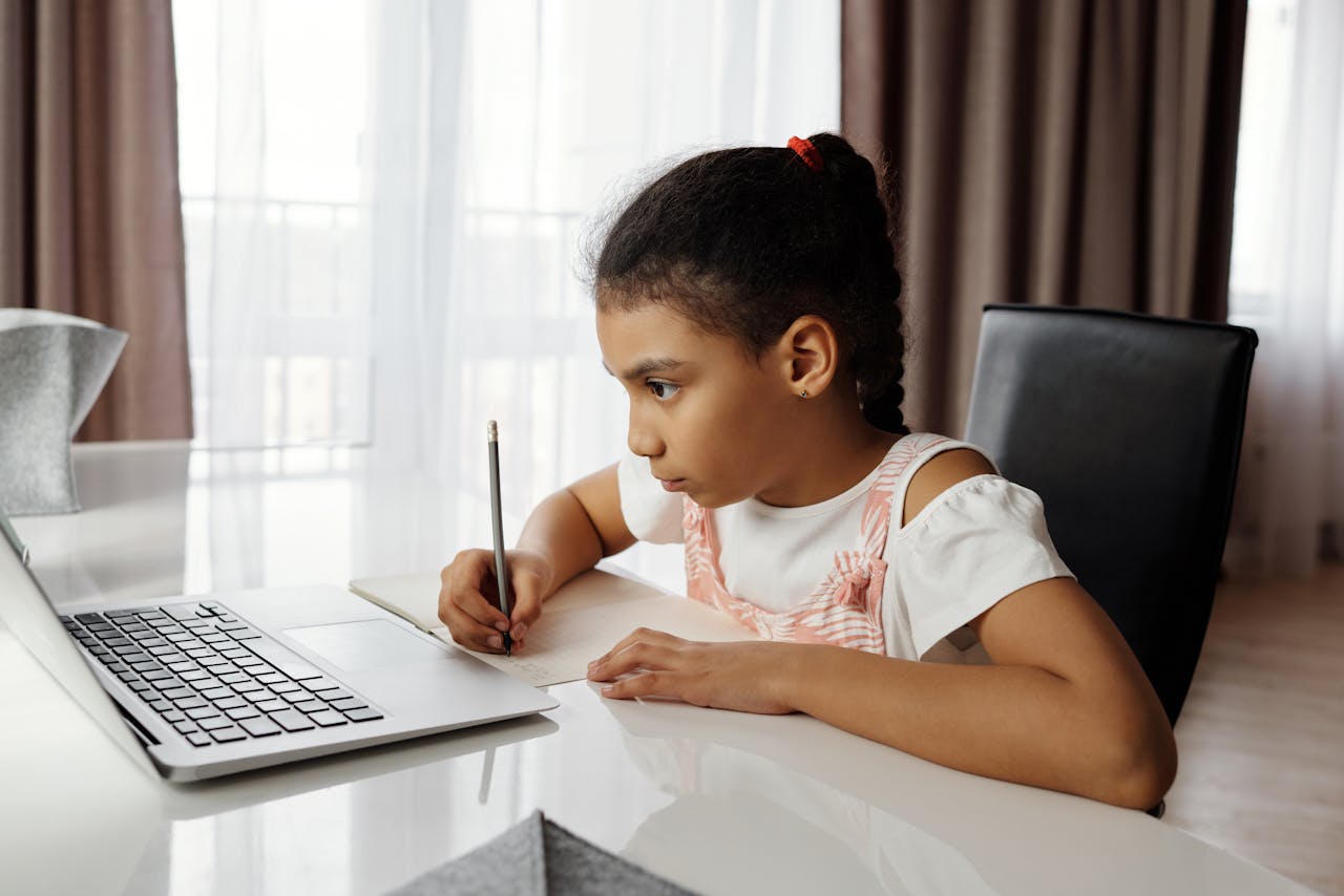 Young girl intensely focused on online learning at home, writing notes by a laptop.